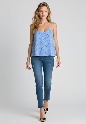 Mujer joven con el pelo rubio largo, que lleva una blusa sin mangas azul claro con lunares, vaqueros ajustados azules y tacones negros abiertos en la punta.