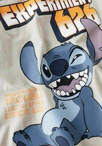 Witte stof met een blauwe cartoonfiguur met grote oren en tanden. De tekst luidt "EXPERIMENT 626" en "PROGRAMMED AS TROUBLE."