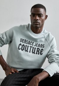 Světle modrá mikina s nápisem "VERSACE JEANS COUTURE" v tučném černém písmu, kulatým výstřihem, žebrovanými manžetami a uvolněným střihem.