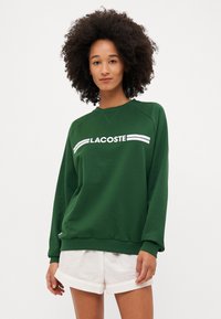 Groene sweater van katoen met een geribde halslijn. Voorzien van een wit "LACOSTE"-logo en twee witte strepen over de borst.