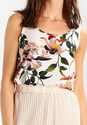 Kvinde iført hvid ærmeløs top med stort blomsterprint og plisseret beige nederdel, vist fra skuldre til talje.