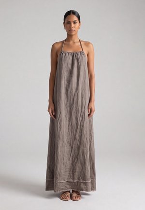 Donna con capelli scuri che indossa un lungo vestito taupe ampio con collo halter e sandali, in piedi davanti a uno sfondo grigio chiaro uniforme.
