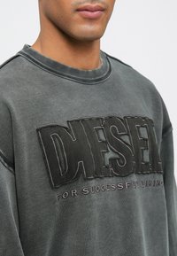 Sweatshirt de algodão cinza escuro com gola canelada, apresentando um logótipo em relevo "DIESEL" e o texto "FOR SUCCESSFUL LIVING" em fio de cor contrastante.