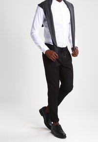 Vit skjorta med knappar, svarta slim-fit byxor och svarta dressade skor. Grå tröja som ligger över axlarna, mjukt tyg och skräddarsydd passform.