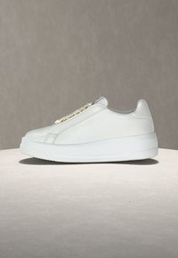 Baskets basses en cuir blanc avec semelle épaisse, œillets dorés et logo discret embossé sur le côté, présentées sur une surface neutre.