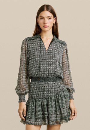 Vestido negro y verde con estampado, mangas largas y transparentes, escote en V, cintura fruncida y dobladillo con volantes. Presenta diseños geométricos.