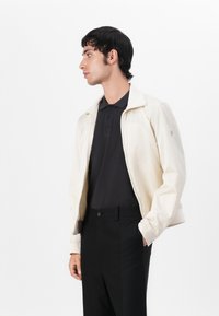 Ljust beige zipjacka med texturerad tyg, med ståkrage och subtil logotypdetalj, buren över en svart polo och svarta byxor.
