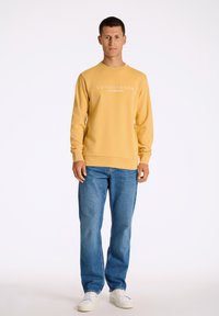 Gul sweatshirt med tryck av "COPENHAGEN" och "LINDBERGH", kombinerad med blå jeans och vita sneakers, stående mot en enfärgad bakgrund.