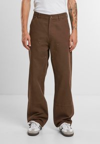 Braune weit geschnittene Hosen aus Baumwolle, mit zwei vorderen Taschen und metallic Akzenten. Kombiniert mit weißen Sneakers.