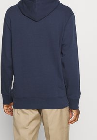 Man med en marinblå hoodie och ljusbruna byxor sedd bakifrån mot en enfärgad ljus bakgrund.