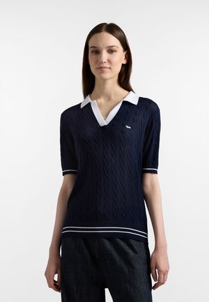 Donna in piedi che indossa un maglione a maniche corte blu navy con lavorazione a trecce, colletto bianco e rifiniture a righe, abbinato a pantaloni scuri.