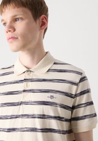 Beige polo triko s námořnickými modrými vodorovnými pruhy, má límec, dva knoflíky a vyšívané logo na hrudi. Hladká textura.