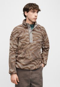 Νέος άνδρας που φοράει καφέ και μπεζ φλις πουλόβερ με μοτίβο και ψηλό γιακά, στέκεται με το ένα χέρι στην τσέπη μπροστά σε απλό φόντο.