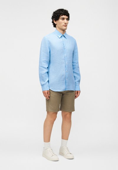 Άνδρας φορώντας ανοιχτό μπλε πουκάμισο με μακριά μανίκια και κουμπιά, χακί σορτς, λευκές κάλτσες και λευκά αθλητικά παπούτσια, στέκεται μπροστά σε απλό φόντο.