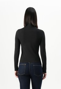 Een persoon met steil donker haar staat met de rug naar ons gekeerd, draagt een aansluitend zwart langemouwen shirt en donkerblauwe jeans met achterzakken.