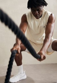 Beige sporttopp, fehér rövidnadrág és fehér edzőcipő. Az illető edzés közben van, fekete csata kötelet tart a kezében.