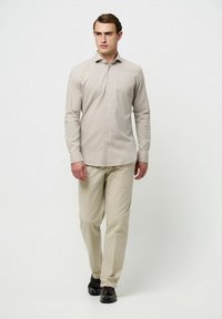 Camisa de botones color beige claro con una textura sutil, combinada con pantalones caqui claros y zapatos negros, en un modelo de pie erguido.