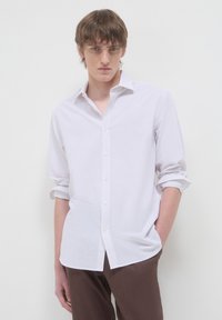 Giovane uomo con capelli castani che indossa una camicia bianca con bottoni e maniche arrotolate, e pantaloni marroni, in piedi davanti a uno sfondo bianco uniforme.