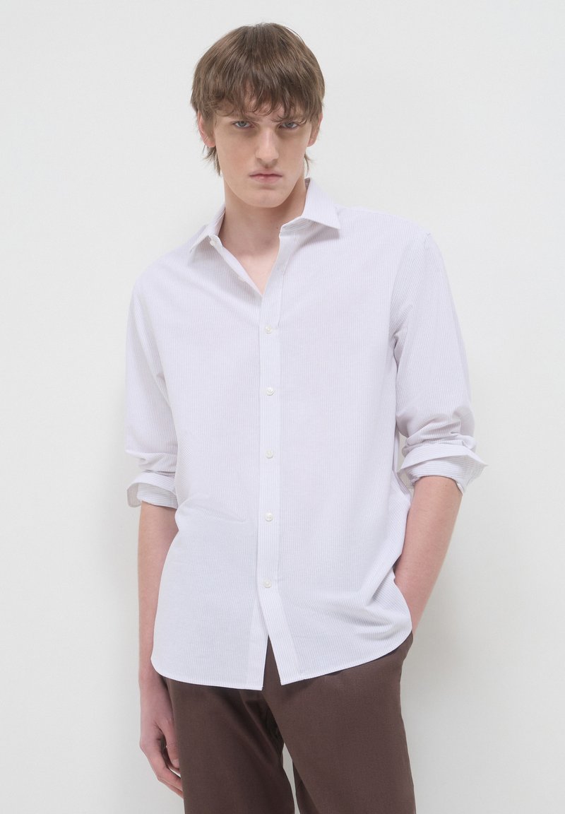 Giovane uomo con capelli castani che indossa una camicia bianca con bottoni e maniche arrotolate, e pantaloni marroni, in piedi davanti a uno sfondo bianco uniforme.