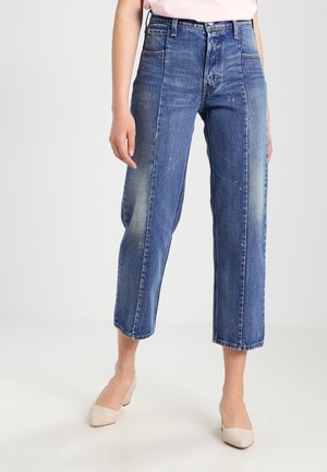 Vrouw draagt verweerde blauwe high-waist cropped jeans met rechte pijpen en beige lage hak schoenen, staand tegen een effen witte achtergrond.