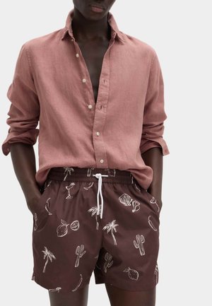 Homme portant une chemise boutonnée rose poussiéreux à manches retroussées et un short marron avec des imprimés tropicaux blancs et d'agrumes.