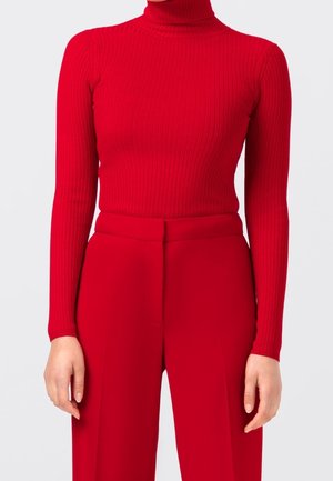 Femme portant un pull à col roulé rouge côtelé ajusté et un pantalon rouge taille haute sur un fond uni.