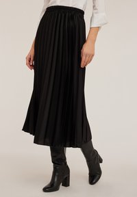 Gonna midi plissettata nera con vita elastica, realizzata in tessuto liscio. Indossata con stivali neri al ginocchio in tessuto texturizzato.