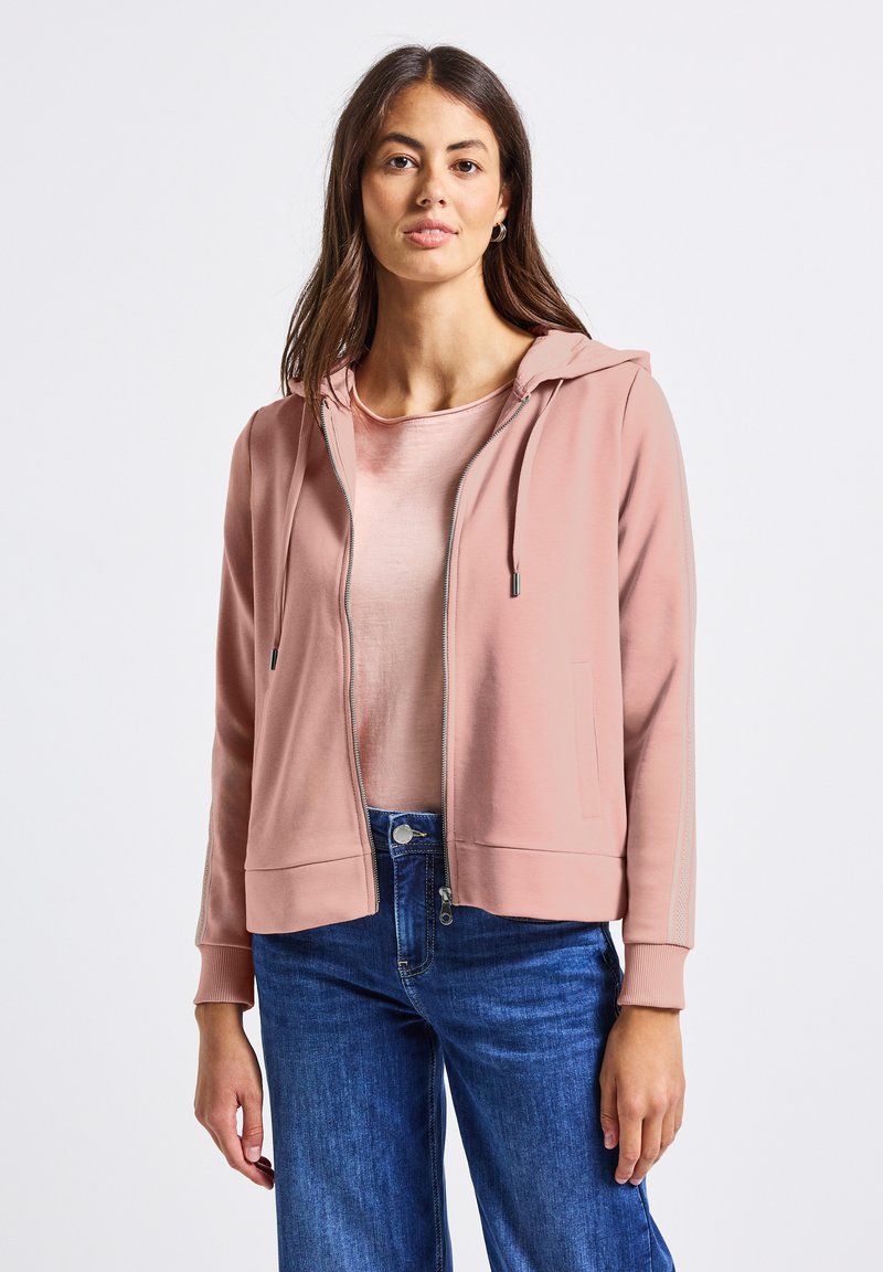 Felpa rosa con zip, cappuccio e tasche frontali, caratterizzata da una texture liscia e strisce chiare a contrasto sulle maniche, indossata sopra una maglietta chiara.
