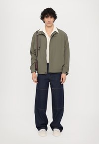 Olivengrønn jakke med krage, lys beige skjorte under, og mørkeblå vide bukser. Hvite sneakers fullfører antrekket.