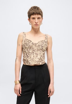 Vrouw met kort haar die een mouwloos topje met luipaardprint draagt, zwarte broek, gouden armband en gouden oorringen, staand tegen een effen achtergrond.