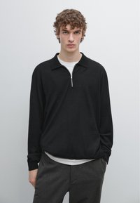 Černý svetr s límcem na zip, vyrobený z měkké tkaniny, nošený přes bílou košili. Model stojí před jednobarevným pozadím.