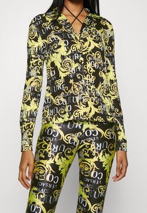 Femme portant un pantalon noir et un haut à manches longues avec des feuilles baroques jaunes et un motif de texte blanc indiquant "Versace Couture"