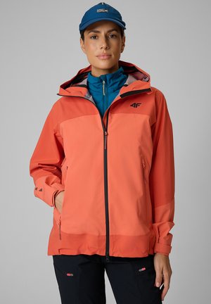 Kobieta w koralowo-pomarańczowej kurtce outdoor z kapturem, niebieskiej warstwie wewnętrznej zapinanej na zamek, czarnych spodniach i niebieskiej czapce, stojąca na szarym tle.