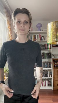 Černé tričko s krátkým rukávem a kulatým výstřihem, zdobené malou nášivkou na hrudi, kombinované s černými kalhotami a světlou dřevěnou podlahou.