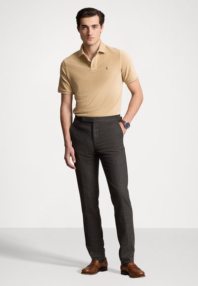 Polo beige con texture a coste e un piccolo emblema, abbinato a pantaloni grigi scuri su misura e scarpe di pelle marrone. Il modello è in piedi.