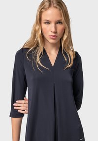 Tunic blu navy con scollo a V e maniche tre quarti. Realizzata in tessuto morbido, con una piega centrale e un piccolo logo decorativo.
