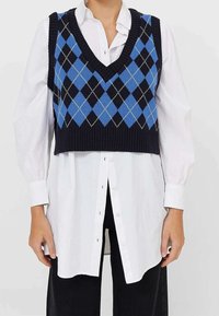 Osoba ubrana w niebieską i czarną kamizelkę w romby, nosząca białą koszulę z długim rękawem i czarne spodnie, stojąca na tle gładkiej ściany.