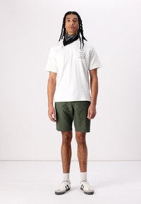 Weißes Baumwoll-T-Shirt mit schwarzem Drucktext, grüne Shorts, weiße Sneaker mit schwarzen Akzenten und ein schwarzes Bandana um den Hals.