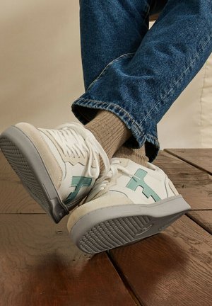 Baskets blanches et beiges avec des accents bleu sarcelle portées avec des chaussettes beiges et un jean bleu, reposant sur un sol en bois.