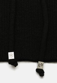 Mustaa neulottua kangasta, jossa on teksturoidut raidoitukset ja kaksi narua hopeanvärisillä neliömetallikoristeilla.