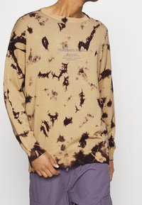 Suéter de punto beige con patrón de tie-dye con acentos marrón oscuro, cuello redondo, mangas largas y estampado gráfico en la parte frontal.