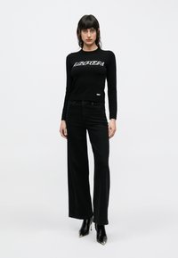 Schwarzes Langarmoberteil mit "ASPEN"-Logo, kombiniert mit hochtaillierten schwarzen Jeans mit weitem Bein. Das Model trägt schwarze spitze High Heels. Minimalistisches Design.