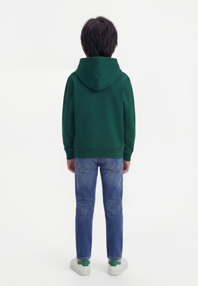 Παιδί που στέκεται γυρισμένο με την πλάτη, φοράει σκουρό πράσινο φούτερ με κουκούλα, μπλε τζιν και άσπρα αθλητικά παπούτσια με πράσινες λεπτομέρειες σε απλό λευκό φόντο.