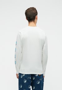 Vīrietis ar muguru, tērpies baltā garajā piedurknē, uz kuras ir zili roku grafiski attēli, kā arī zilās bikses ar baltajiem yin-yang simboliem.