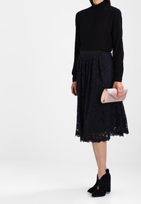 Svart blus med hög krage i kombination med en mörk spetskjol. Modellen håller en rosa kuvertväska. Svarta ankboots kompletterar outfiten.