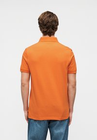 Πορτοκαλ ί πόλο πουκάμισο με κλασικό γιακά, κοντά μανίκια και λεία υφή από βαμβάκι. Συνδυάζεται με μπλε τζιν παντελόνι.