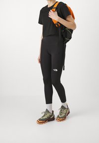 Černé oříznuté tričko, černé legíny s vysokým pasem a bílým logem, šedé ponožky a zeleno-černo-oranžové turistické boty s texturovanými podrážkami.