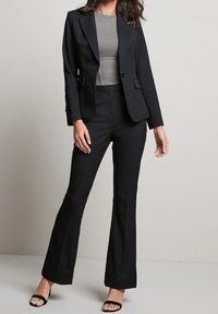 Schwarzer Blazer und passende ausgestellte Hose, figurbetontes Design, glatter Stoff, graues geripptes Top darunter, getragen mit schwarzen Riemchen-High Heels.