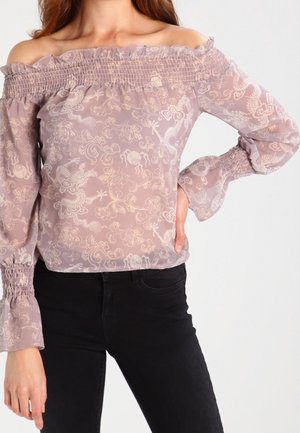 Kobieta w przezroczystej, lawendowej bluzce z odsłoniętymi ramionami, marszczonym dekoltem i mankietami, w połączeniu z czarnymi spodniami z wysokim stanem.