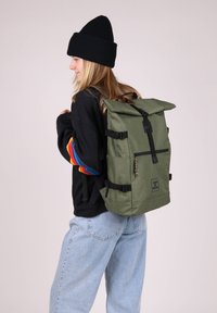 Zaino in tela verde con design a rullo, accessori neri, spallacci regolabili e tasca frontale con zip. Il modello indossa un maglione nero e jeans.